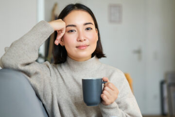 Smiling woman sitting on sofa with her mug, drinking coffee at home and relaxing after work, looking calm and cozy.