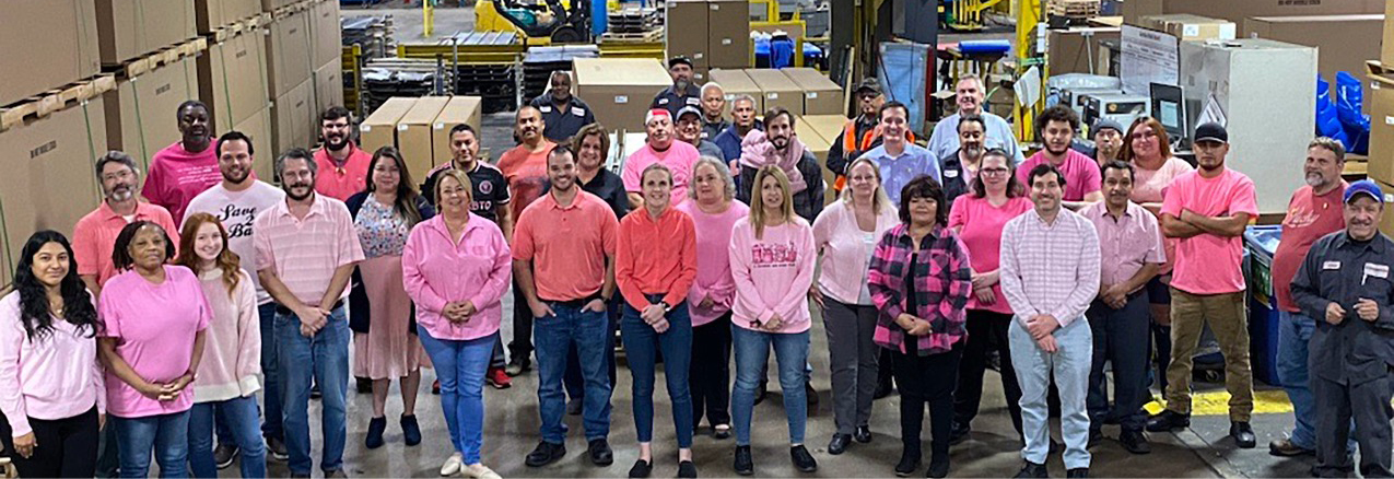 Large group of manufacturing workers all dressed in pink for Breast Cancer Awareness week smiling at camera.