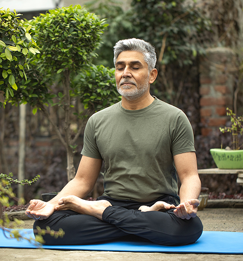 senior man meditating in lotus position while sitting on exercise mat surrounded with plants in park