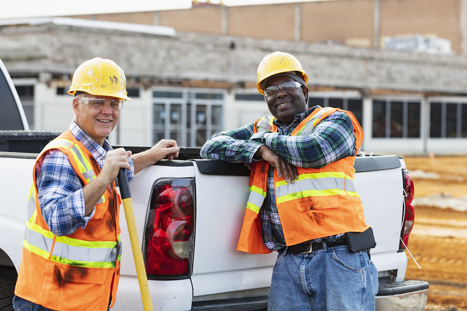 Two multiracial construction workers standing behind a pickup truck. The African-American man, in his 40s, is leaning against the tailgate with his arms crossed. His coworker, a senior man in his 60s, is holding a shovel. They are smiling at the camera.